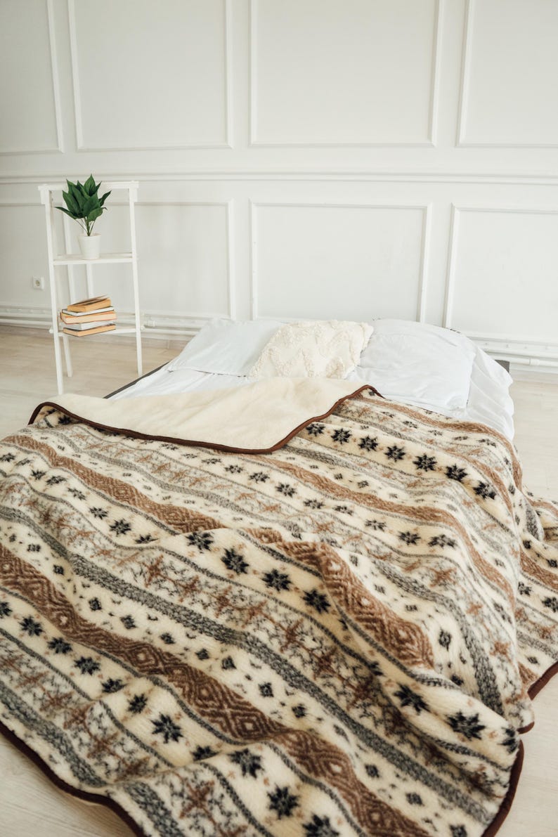 May include: A beige and brown patterned blanket with a dark brown border is draped over a bed. The blanket features a repeating pattern of stripes, snowflakes, and geometric shapes. The bed has white sheets and pillows.