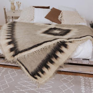 May include: A patterned blanket in neutral tones with a geometric design is draped over a bed. The blanket has a central diamond shape and black borders. The bed is made from wooden pallets, and the room has a neutral colour scheme.