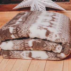 以下が含まれることがあります： 茶色と白の模様の折り畳まれた毛布。毛布は柔らかく、質感のある外観で、きちんと折りたたまれています。模様は繰り返しのデザインのようです。毛布は木製の表面に置かれています。