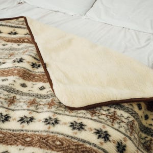 May include: A cozy blanket with a cream-colored, plush interior and a patterned exterior. The exterior features a repeating design in shades of brown, gray, and black. The blanket is placed on a bed with white sheets and pillows.