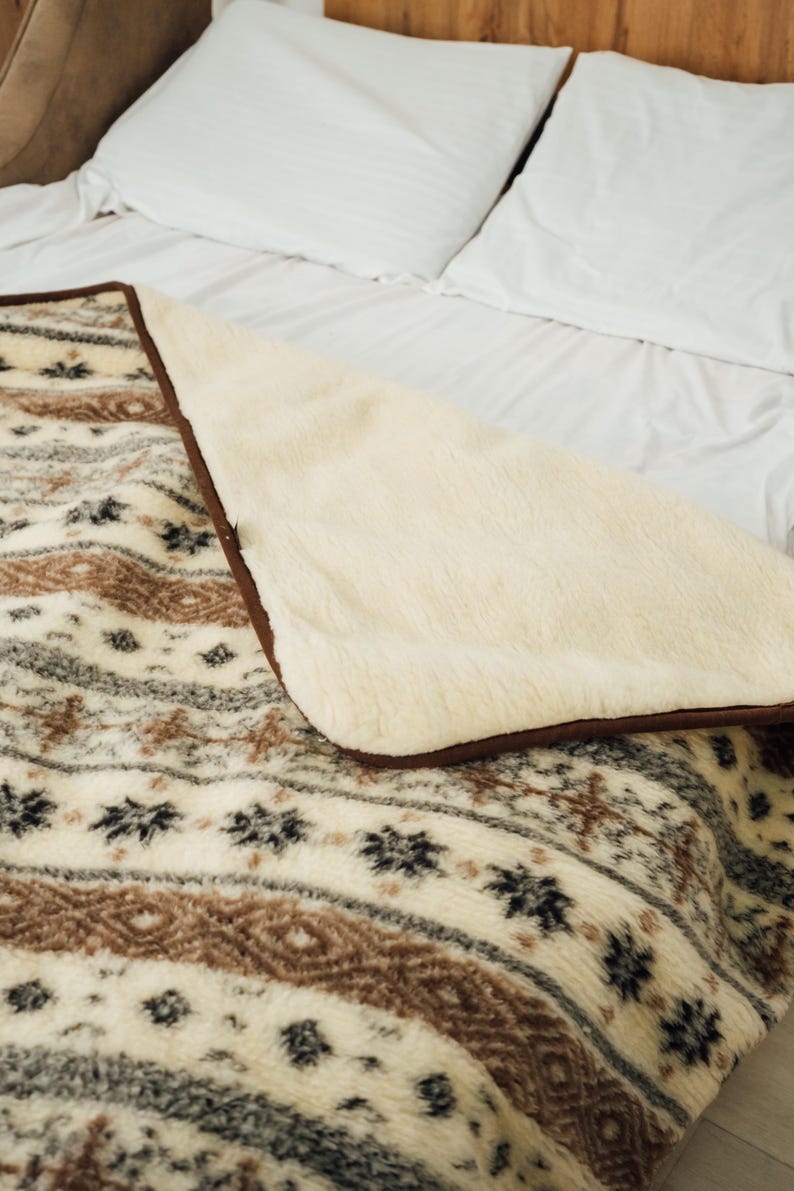 May include: A cozy blanket with a cream-colored, plush interior and a patterned exterior. The exterior features a repeating design in shades of brown, gray, and black. The blanket is placed on a bed with white sheets and pillows.