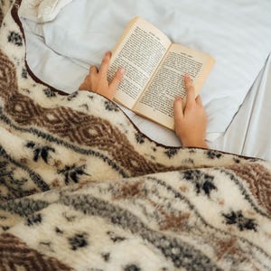 May include: A cozy, patterned blanket in cream, brown, and gray covers a bed. The blanket features a geometric design. A person is reading a book with aged pages, resting on the bed. The scene evokes comfort and relaxation.