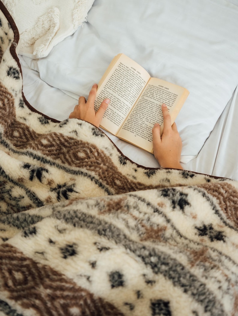 May include: A cozy, patterned blanket in cream, brown, and gray covers a bed. The blanket features a geometric design. A person is reading a book with aged pages, resting on the bed. The scene evokes comfort and relaxation.