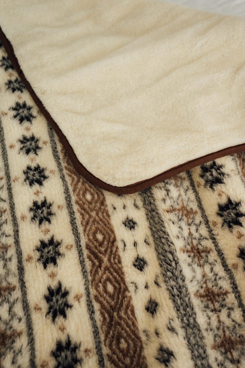 May include: A cozy, cream-colored blanket with a soft, plush texture. The blanket features a patterned design with brown, black, and gray geometric shapes and stripes. The edge is trimmed with a brown border.