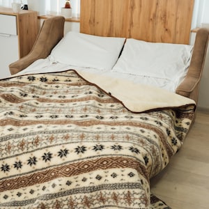 May include: A bed with white pillows and a patterned blanket. The blanket features a cream, brown, and black geometric design. The bed has a wooden headboard and a neutral-toned upholstered frame.