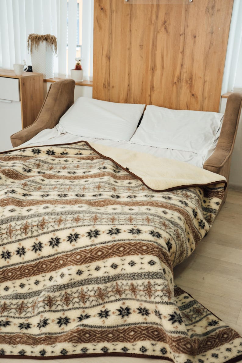 May include: A bed with white pillows and a patterned blanket. The blanket features a cream, brown, and black geometric design. The bed has a wooden headboard and a neutral-toned upholstered frame.