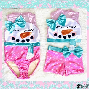 May include: A pink and white snowman-themed swimsuit and shorts set. The swimsuit and shorts feature a pink snowflake pattern. The snowman design includes a teal bow, orange nose, and black eyes. The set is designed for children.
