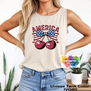 May include: Ivory unisex tank top featuring a graphic design with the word "AMERICA" in red, white, and blue. The design includes a bow with a stars and stripes pattern and two cherries. The tank top is made of a soft material.
