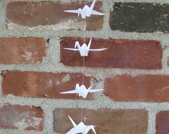 Może przedstawiać: Sznur białych żurawi origami wiszący na ceglanej ścianie. Żurawie są połączone białą nicią i rozmieszczone w równych odstępach.
