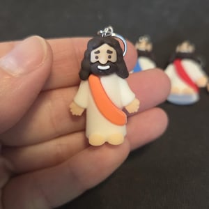 Może przedstawiać: Mała figurka Jezusa Chrystusa, z białą szatą, pomarańczową szarfą oraz ciemnymi włosami i brodą. Figurka trzymana jest w dłoni, a w tle widoczne są inne podobne figurki. Figurka posiada srebrną pętlę do zawieszenia.