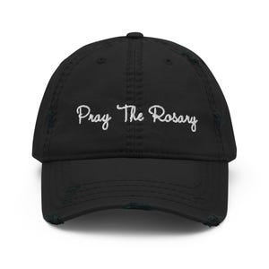 Könnte beinhalten: Eine schwarze Baseballkappe mit einem abgenutzten, distressed Finish an Schirm und Krone. Auf der Vorderseite befindet sich der weiß gestickte Schriftzug 'Pray The Rosary' in Schreibschrift. Die Kappe hat ein strukturiertes Vorderteil.