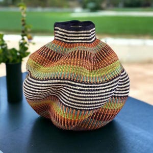 May include: A decorative, handwoven basket with a distinctive, undulating form. The basket displays a blend of colours: brown, orange, green, and blue, with horizontal black and white stripes around the neck. It is placed on a dark surface.