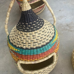 Puede incluir: Cesta tejida a mano con una forma única y un asa. La cesta presenta una parte superior negra, una franja colorida de color turquesa, amarillo y rojo, y una base tejida natural. La cesta tiene una abertura lateral.