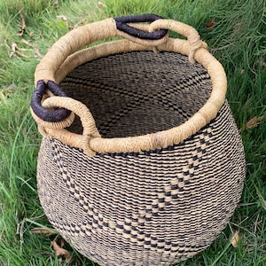 May include: A round woven basket with a natural tan and black pattern. The basket has a sturdy, looped handle with dark brown accents. The basket is sitting on a bed of green grass.