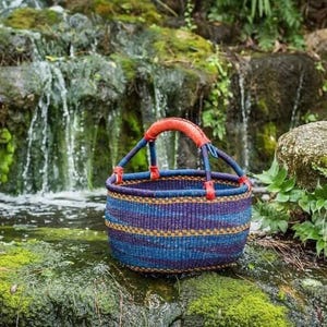 Peut inclure: Un panier rond tissé avec des rayures horizontales bleues, violettes et dorées. Le panier a deux anses bleues avec des accents en cuir rouge. Le panier est posé sur un rocher recouvert de mousse près d'une cascade.