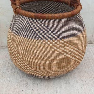 Puede incluir: Cesta tejida a mano con forma redondeada y asa resistente. La cesta presenta un diseño de dos tonos, con un patrón tejido en beige y negro en la mitad inferior y un borde marrón. El asa está envuelta en un material de color más claro.