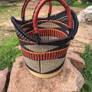 Peut inclure: Panier tissé avec un motif géométrique noir, orange et beige. Le panier a deux poignées arquées enveloppées de cuir marron. Le panier est posé sur un gros rocher rugueux brun rougeâtre.