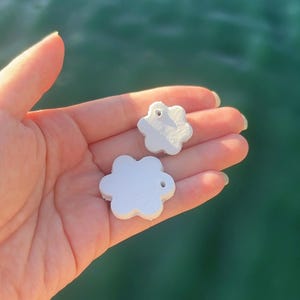 May include: Two small, white, flower-shaped charms with a hole for stringing. The charms are held in a hand, with a blurred background of water. The charms have a smooth, matte finish and are likely made of plastic or resin.