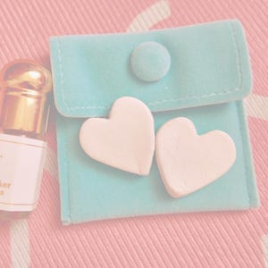 May include: A small perfume bottle with a gold cap and a label, next to a light blue pouch with two white heart-shaped decorations. The items are on a pink surface.
