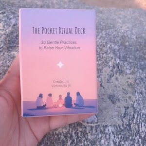 May include: A card deck titled "The Pocket Ritual Deck" with the text "30 Gentle Practices to Raise Your Vibration." The box has a pastel pink and purple gradient background with a star and a group of people around a fire.