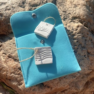 May include: Two small, square, white pendants with a textured design, each suspended by a natural twine loop, rest on a turquoise blue pouch. The pendants are likely made of a stone-like material. The pouch is closed with a snap.