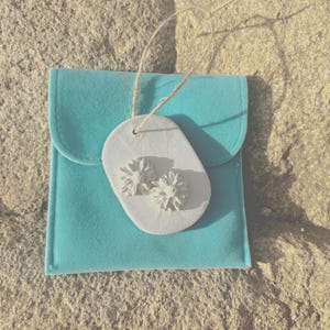May include: A turquoise pouch with a white tag adorned with two white, flower-shaped decorations. A natural-coloured string attaches the tag to the pouch. The background is a textured, light brown surface.