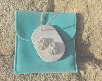 May include: A light blue pouch with a white tag featuring two white flower-shaped embellishments. The tag is attached to the pouch with a natural-colored string. The background is a textured, light brown surface.