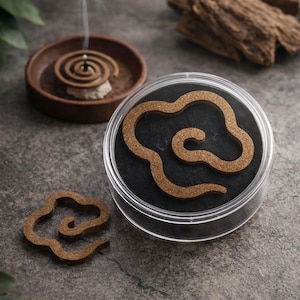 May include: Aromatic incense spirals and wood chips are displayed on a gray surface. One spiral is burning, emitting smoke. A container holds a cloud-shaped incense, and another sits separately. The scene suggests relaxation and aromatherapy.