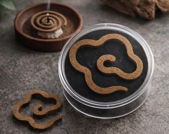 May include: Aromatic incense spirals and wood chips are displayed on a gray surface. One spiral is burning, emitting smoke. A container holds a cloud-shaped incense, and another sits separately. The scene suggests relaxation and aromatherapy.