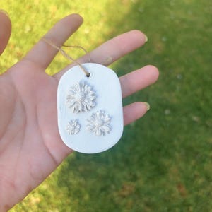 May include: A white, oval-shaped ornament with three embossed flower designs. The ornament is suspended by a thin, natural-colored twine. The background is a blurred view of green grass.