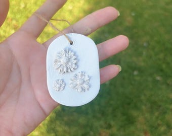 May include: A white, oval-shaped ornament with three embossed flower designs. The ornament is suspended by a thin, natural-colored twine. The background is a blurred view of green grass.