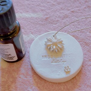 May include: A brown essential oil bottle next to a white, circular aromatherapy disc. The disc has a daisy design and the word "Inspiration" embossed on it. A string is attached for hanging.
