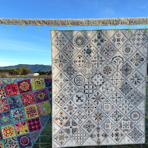 May include: Two quilts hanging on a washing line against a blue sky. The quilt on the left is a patchwork of colourful squares with geometric designs. The quilt on the right is a white quilt with a pattern of small, intricate designs in shades of grey, brown, and blue.