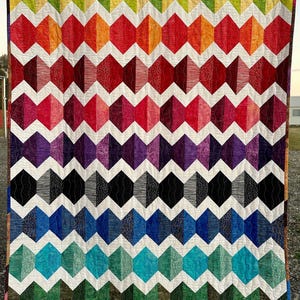 May include: A colorful handmade quilt with a geometric design. The quilt features rows of diamond shapes in a spectrum of colors, including red, orange, purple, blue, and green, separated by white spaces. The quilt is hanging outdoors.