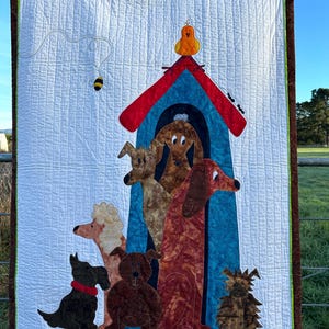 Può includere: Un arazzo da parete trapuntato colorato con una cuccia per cani blu con tetto rosso. Un uccello giallo si siede sulla cima della casa. Cinque cani di razze e colori diversi stanno davanti alla casa. Un'ape e le formiche sono anche raffigurate sullo sfondo bianco.
