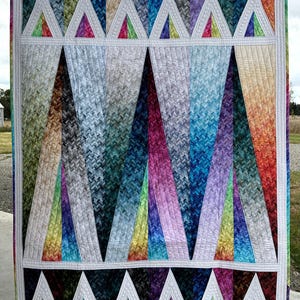Op de afbeelding: Een kleurrijk quilt met een geometrisch patroon van driehoeken. De quilt is gemaakt van stof met een chevron-patroon en is gequilt met wit garen. De quilt hangt aan een waslijn.