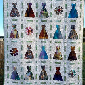 May include: A white quilt with a green grid pattern. The quilt is decorated with 18 appliqued fabric dresses in various colours and patterns. There are also 5 appliqued fabric flowers in the centre of each row.