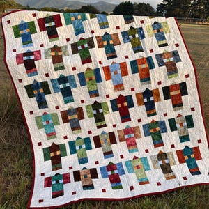 May include: A white quilt with a red border. The quilt is made up of squares with a pattern of colourful geometric shapes. The quilt is laid out on a grassy field.
