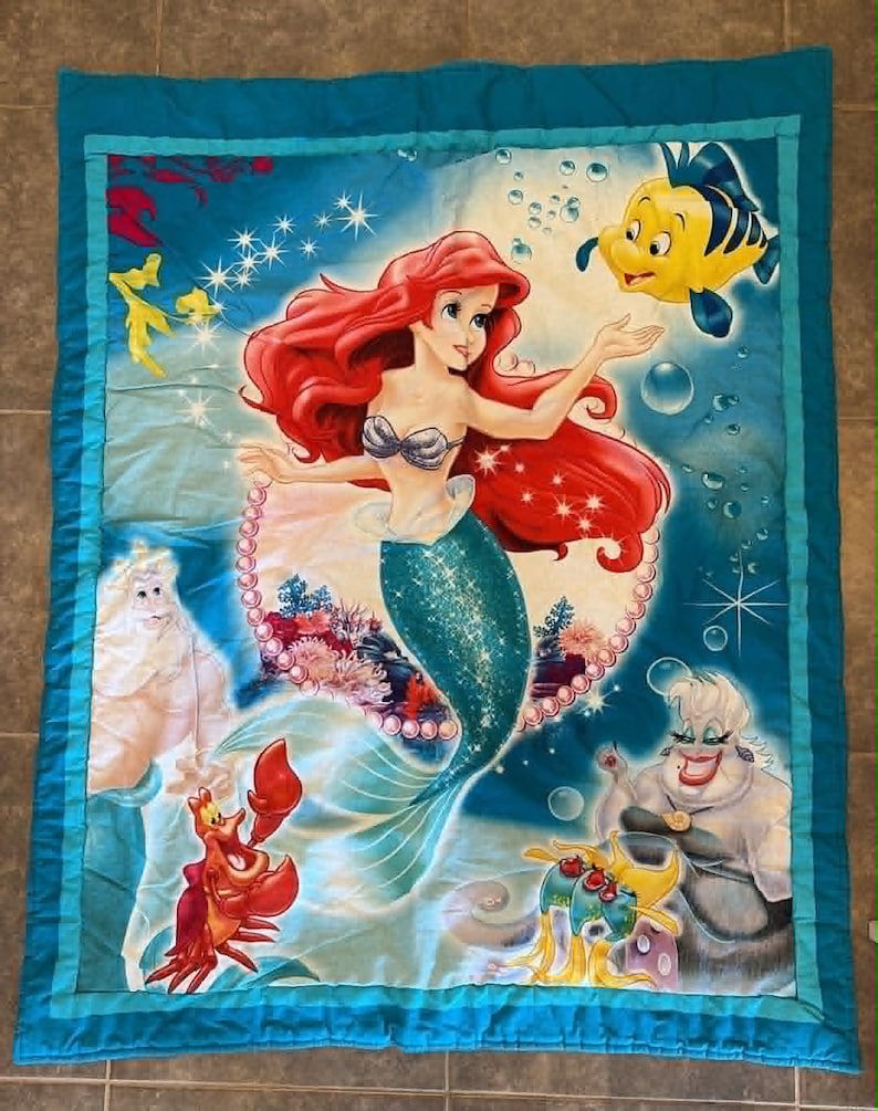 May include: A rectangular quilt featuring a vibrant underwater scene with Ariel the mermaid, Flounder the yellow fish, Sebastian the red crab, King Triton, and Ursula. The design includes blue water, bubbles, and a pearl border on a teal background.