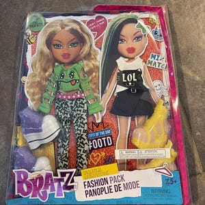 Peut inclure: Un coffret Bratz Fashion Pack avec deux poupées aux tenues stylées. L'une a les cheveux blonds et un haut vert, l'autre a les cheveux verts et un haut avec l'inscription "LOL". L'emballage comprend le texte "Fashion Pack" et "Deluxe Luxeuse".