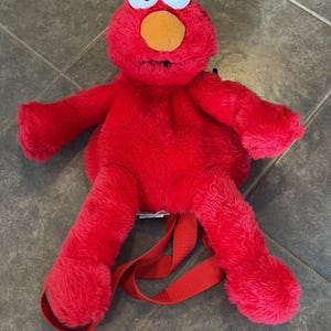 Op de afbeelding: Een felrode pluche rugzak in de vorm van het personage Elmo. De tas heeft een pluizige rode buitenkant, twee grote witte plastic ogen met zwarte pupillen en een oranje neus. Het bevat verstelbare rode stoffen banden aan de achterkant.