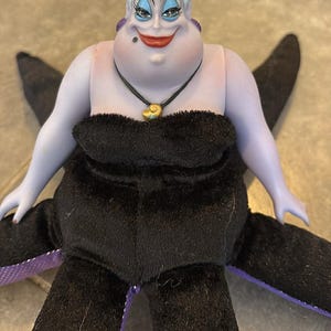 May include: A doll of Ursula, the sea witch from Disney's The Little Mermaid. The doll has a purple face and arms, white hair, and a black dress. She wears a necklace with a gold shell pendant and has black tentacles.