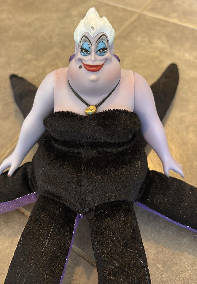 May include: A doll of Ursula, the sea witch from Disney's The Little Mermaid. The doll has a purple face and arms, white hair, and a black dress. She wears a necklace with a gold shell pendant and has black tentacles.