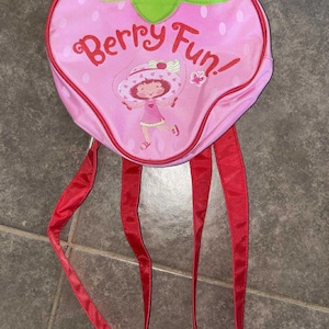May include: A pink strawberry-shaped backpack with red straps and a green leaf-shaped top. The backpack features a cartoon illustration of a girl jumping rope and the words "Berry Fun!"