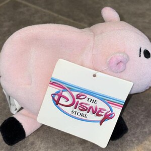 Puede incluir: Un peluche de cerdo rosa con pezu&ntilde;as negras, una cola rizada y un hocico rosa. El cerdo tiene ojos negros y una etiqueta que dice "The Disney Store" en rosa y azul.