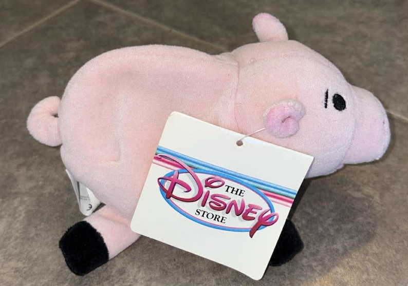Puede incluir: Un peluche de cerdo rosa con pezu&ntilde;as negras, una cola rizada y un hocico rosa. El cerdo tiene ojos negros y una etiqueta que dice "The Disney Store" en rosa y azul.