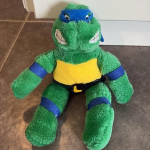 Peut inclure: Une peluche de Leonardo des Tortues Ninja. La figurine verte porte un masque bleu, des bandes bleues aux bras et aux jambes, un plastron jaune et une ceinture noire avec une boucle "L" jaune. Elle a des dents blanches et est en mati&egrave;re douce et duveteuse.