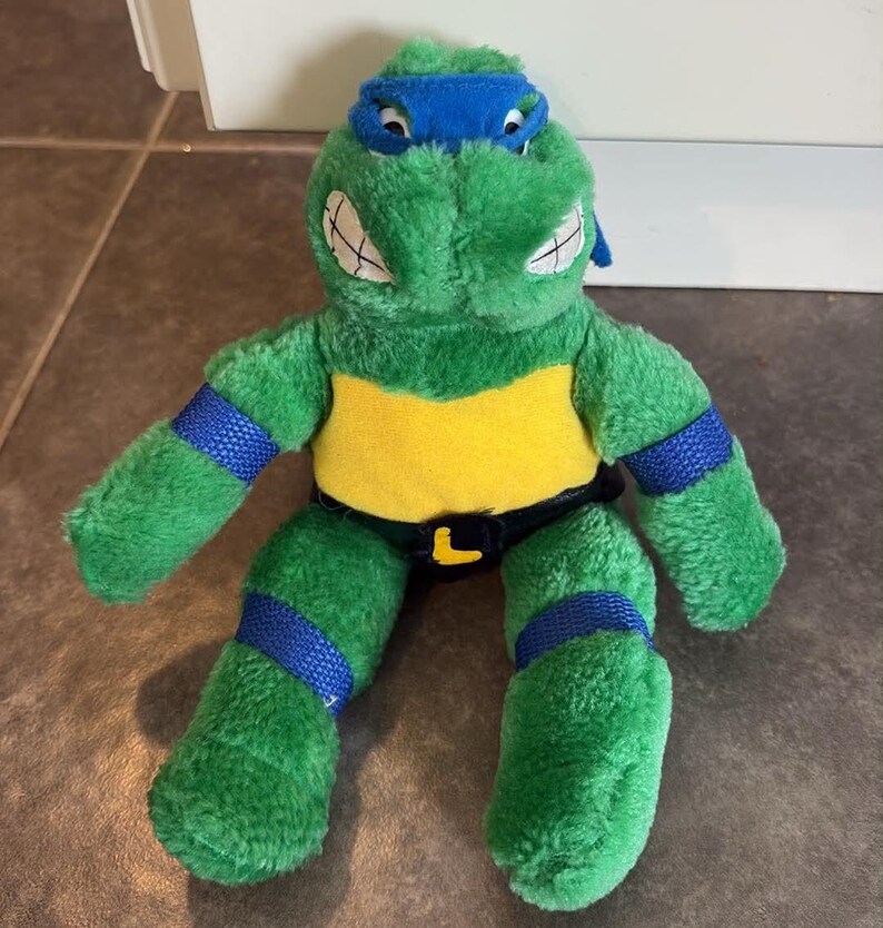 Peut inclure: Une peluche de Leonardo des Tortues Ninja. La figurine verte porte un masque bleu, des bandes bleues aux bras et aux jambes, un plastron jaune et une ceinture noire avec une boucle "L" jaune. Elle a des dents blanches et est en mati&egrave;re douce et duveteuse.