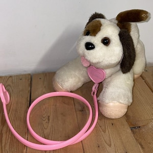 Puede incluir: Un perro de peluche con pelaje blanco, orejas y manchas marrones, y una etiqueta rosa en forma de corazón. Tiene una correa rosa. El juguete está diseñado para parecerse a un cachorro.