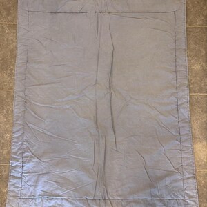 May include: A rectangular, light gray fabric blanket featuring a wide border stitched around the perimeter. The textile has a soft, slightly wrinkled texture and includes small metal grommets at the top two corners for hanging or display purposes.